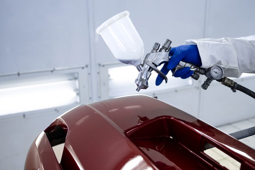 Car painter in protective clothes and mask painting automobile bumper with metallic paint and varnish in chamber workshop.